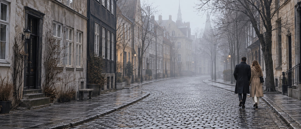 A quiet cobblestone street in soft morning fog, two figures walking away in coats