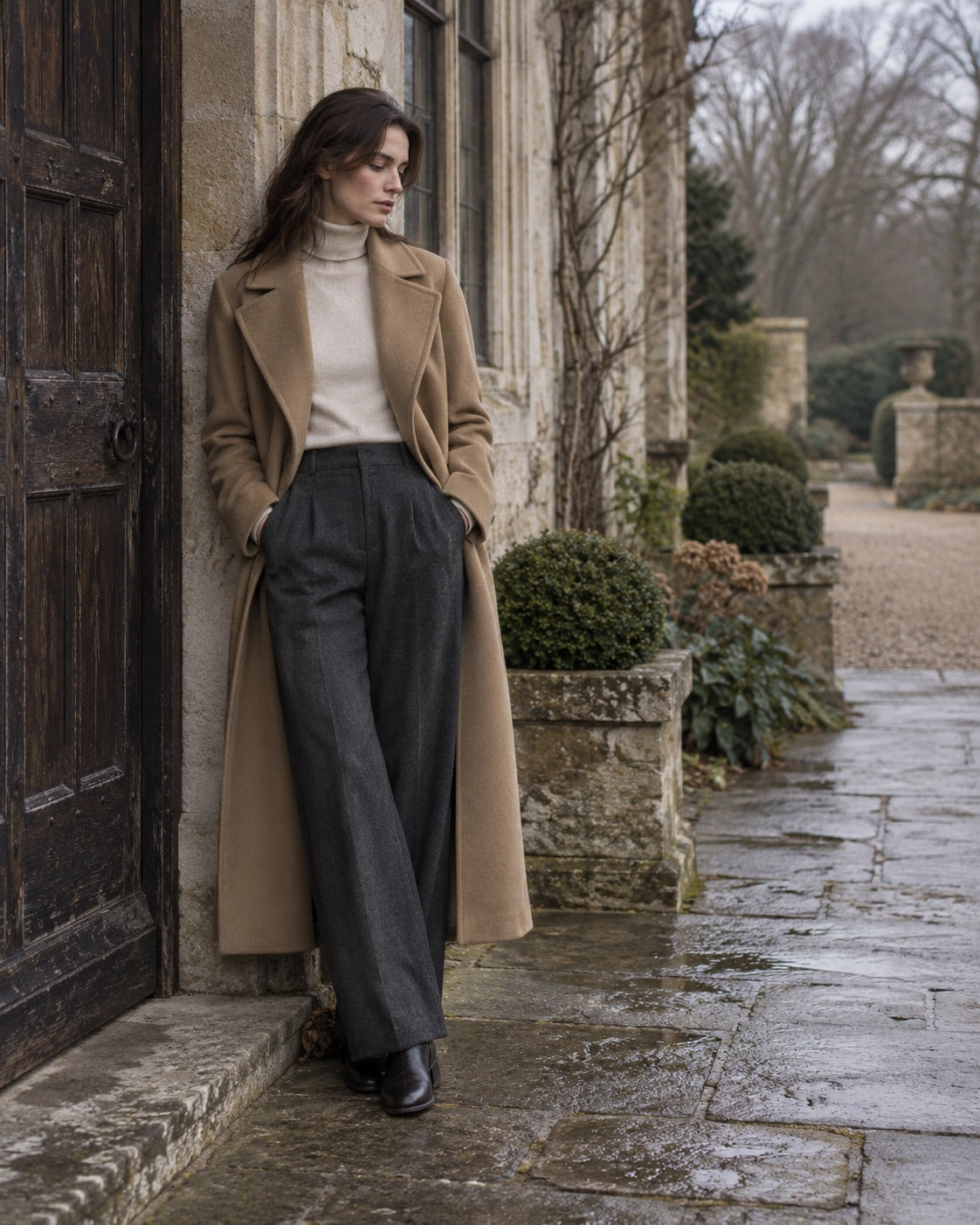 A woman in a camel coat leaning against a stone building in a quiet courtyard